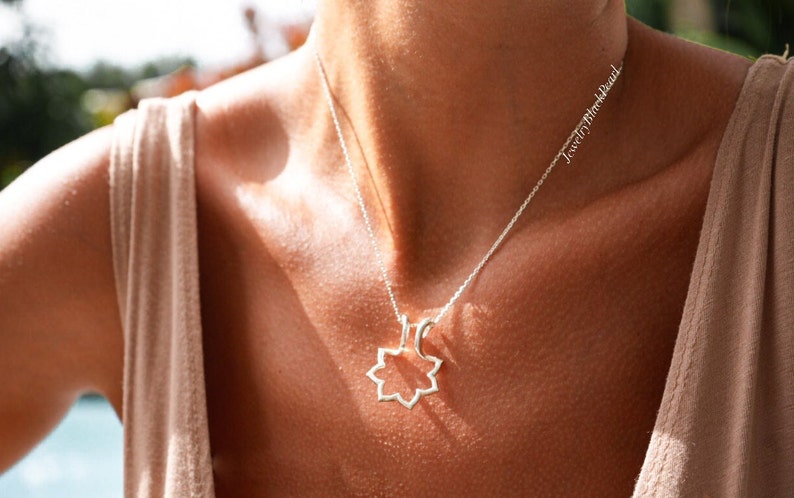 May include: A silver necklace with a geometric pendant in the shape of an eight-pointed star. The necklace is worn on a person with fair skin, and the chain is delicate. The text "JewelryBlackPearl" is visible on the chain.
