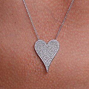 May include: A silver chain necklace with a large, heart-shaped pendant covered in small, clear cubic zirconia stones.