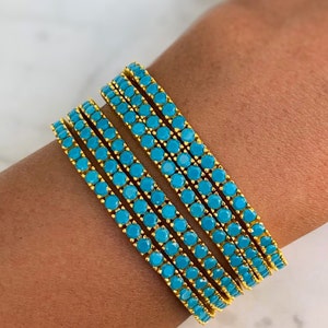 May include: Set of five gold bangles with turquoise blue stones. The bangles are stacked on a person's wrist.