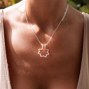 May include: A silver necklace with a geometric floral pendant. The pendant is a stylized openwork design, suspended from a delicate chain. The necklace is worn against a neutral-toned top, with the text "jewelrybyblackpearl" visible.