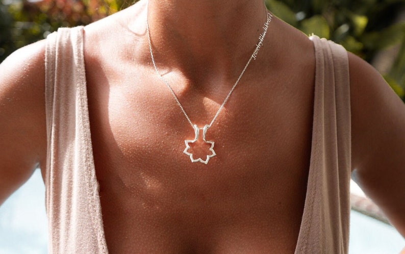 May include: A silver necklace with a geometric floral pendant. The pendant is a stylized openwork design, suspended from a delicate chain. The necklace is worn against a neutral-toned top, with the text "jewelrybyblackpearl" visible.