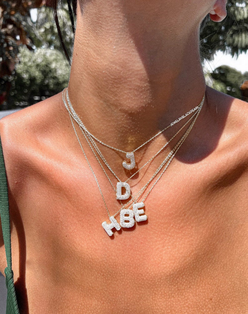 Puede incluir: Primer plano del pecho de una mujer que lleva un collar de capas con letras engastadas en diamantes que deletrean "HBE" y "JD".