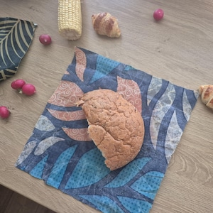 Puede incluir: Un bollo de pan integral se encuentra sobre un envoltorio de alimentos reutilizable de cera de abeja con un diseño de hojas azules, turquesas y naranjas. Otros alimentos incluyen un cruasán, mazorca de maíz y rábanos. El envoltorio reemplaza el film plástico.