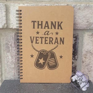 May include: A spiral-bound notebook with a tan cover featuring dark brown text that reads THANK a VETERAN. The design includes a graphic of two military dog tags on a chain, flanked by two small stars, centered below the printed text.