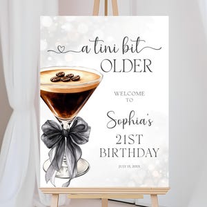 Puede incluir: Un letrero blanco sobre un caballete con el texto "a tini bit OLDER" y "Welcome to Sophia's 21st Birthday". El letrero presenta una ilustración de acuarela de un cóctel de café con un lazo negro.