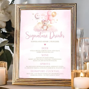 May include: A framed pink and gold sign with the text "Signature Drinks" and a teddy bear illustration. The sign lists beer, wine, spirits, and a signature cocktail. The sign is surrounded by candles and flowers.