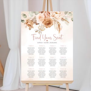 Könnte beinhalten: Ein Hochzeits-Sitzplan auf einer Staffelei. Das Schild zeigt oben ein Blumendesign in Beige- und Brauntönen mit den Worten "Find Your Seat" in eleganter Schrift. Die Namen der Gäste sind unter den Tischnummern aufgeführt.