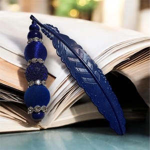 Puede incluir: Un marcapáginas de pluma azul con un borla de cuentas. La borla está hecha de terciopelo azul, cuentas azules y cuentas plateadas.
