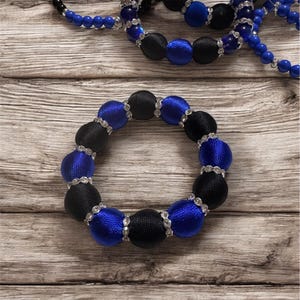 Puede incluir: Un brazalete de cuentas azul y negro con detalles plateados. El brazalete está hecho de tela y tiene una forma circular.