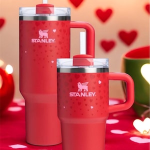 May include: Two red Stanley travel mugs with a heart pattern and the Stanley logo. The larger mug has a straw and a handle. The smaller mug has a handle.