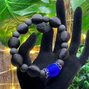Puede incluir: Una pulsera de cuentas marrón oscuro con una pieza central azul vibrante. La pulsera se muestra en una mano negra, mostrando la textura y forma natural de las cuentas. Las cuentas parecen ser de madera o un material similar.