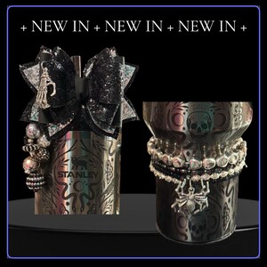 May include: Two decorated Stanley tumblers. One has a black bow and a skeleton charm. The other has a spider charm and skull and moon designs. Both are adorned with beaded bracelets. The text "+ NEW IN + NEW IN + NEW IN +" is at the top.