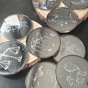 Puede incluir: Colección de posavasos redondos de color plateado con diseños grabados de constelaciones del zodíaco. Los posavasos están dispuestos sobre rodajas de madera, mostrando símbolos astrológicos como Libra, Piscis y Leo. Las superficies reflectantes crean un efecto de espejo.