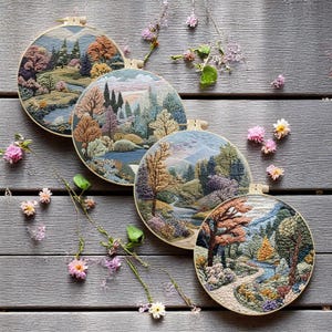 May include: Four embroidered hoop art pieces depicting scenic landscapes with trees, rivers, and mountains. The hoops are arranged in a row on a wooden surface with pink flowers scattered around.
