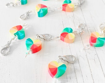 Può includere: Un set di marcatori di punto a forma di cuore arcobaleno con chiusure argentate e perline trasparenti. I marcatori sono realizzati in pasta polimerica e presentano un motivo a spirale.