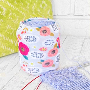May include: A light blue yarn holder with a floral pattern and the text "fresh out of fucks". The holder is filled with light blue yarn. Knitting needles and a partially knitted project are in the foreground.
