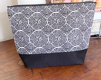 Pode incluir: Uma bolsa com zíper estampada em preto e branco. A parte superior apresenta um design circular repetitivo, enquanto a parte inferior é preta lisa. A bolsa está em uma superfície de madeira.