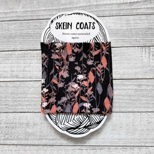 May include: A black fabric skein coat with a floral pattern in shades of pink and gray. The fabric is wrapped around a white card with the text "Skein Coats" and "Never come unraveled again."