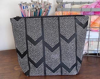 Pode incluir: Bolsa com fecho de correr com padrão chevron preto e branco. A bolsa está sobre uma superfície de madeira com agulhas de tricô no fundo.