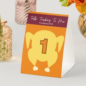 May include: Orange and white table number card with a cartoon turkey illustration and the text "Talk Turkey To Me" and "Friendsgiving 2024". The number "1" is in a brown circle on the turkey.