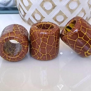 Pode incluir: Três rolhas decorativas para garrafas com um padrão de girafa em tons de castanho e amarelo. As rolhas são cilíndricas e têm um orifício no centro. O fundo inclui uma superfície branca e um objeto de vidro decorativo.