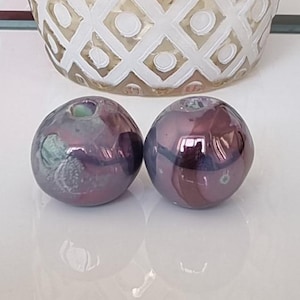 Pode incluir: Duas contas de vidro iridescentes com um esquema de cores roxo e verde. As contas são redondas e têm um acabamento liso e brilhante.