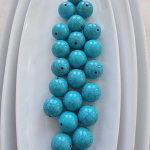 Puede incluir: Un conjunto de cuentas redondas turquesas con un patrón agrietado en forma de telaraña. Las cuentas están dispuestas sobre una superficie blanca y curvada, creando una presentación visualmente atractiva. Ideales para la creación de joyas y proyectos de artesanía.