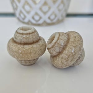 Könnte beinhalten: Zwei pilzförmige Flaschenverschlüsse mit Craquelé-Glasur. Die Verschlüsse sind beige und haben eine abgerundete Form. Der Hintergrund enthält ein dekoratives Glasobjekt.