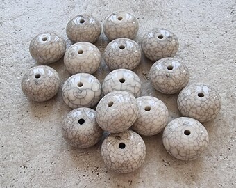 Könnte beinhalten: Eine Sammlung von 16 weißen Keramikperlen mit einer craquelé-Glasur. Die Perlen sind rund und haben ein Loch in der Mitte.