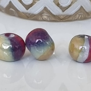 Puede incluir: Tres cuentas de vidrio redondas con un patrón de remolino multicolor. Las cuentas son amarillas, blancas, moradas y rojas.