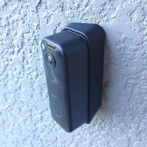 May include: A black rectangular doorbell camera mounted on a textured white wall. The camera has a lens, a light, and a button. The doorbell is designed for home security and smart home integration.