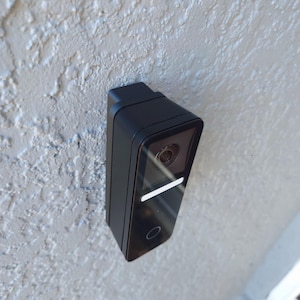 Puede incluir: Un timbre inteligente negro y rectangular montado en una pared texturizada de color gris claro. El timbre tiene una lente de cámara, una luz blanca horizontal y un botón circular en la parte inferior.