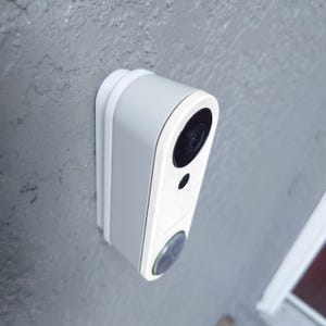 May include: A white and gray smart doorbell mounted on a textured gray wall. The doorbell has a camera lens, a small black sensor, and a circular button. The device is designed with rounded edges and a sleek, modern aesthetic.