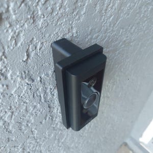 May include: A black security camera fixed to a textured, light grey wall. The camera features a lens and a button, with the brand name "ring" visible. The camera is rectangular and has a modern design. The wall has a rough, stucco-like finish.