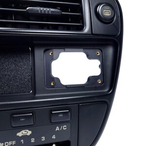 May include: Black plastic car dashboard with a blank rectangular cutout for a radio or other device. The dashboard has a few buttons and a small air vent.