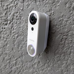 May include: A white SimpliSafe video doorbell with a silver button and a black camera lens. The doorbell is mounted on a gray stucco wall.