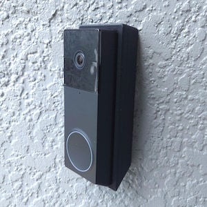 Może przedstawiać: Czarny inteligentny dzwonek do drzwi z kamerą i okrągłym światłem, zamontowany na teksturowanej białej ścianie. Dzwonek ma elegancki, nowoczesny design, z prostokątnym kształtem i wyraźnym obiektywem.