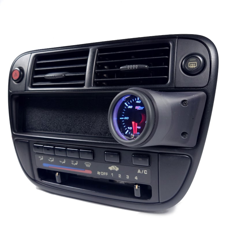 May include: A black car dashboard with a blue and red gauge mounted on the right side. The gauge has a white needle and a black face with white numbers. The dashboard also has a black radio and climate control panel with buttons and knobs.