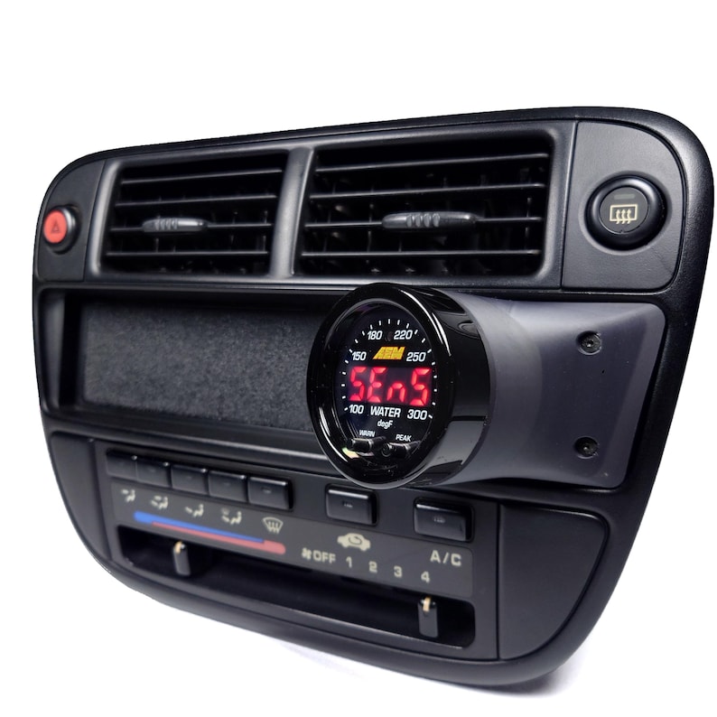 May include: A black car dashboard with a digital water temperature gauge mounted to the right of the air vents. The gauge reads "SE05" and has a range of 100 to 300 degrees Fahrenheit. The gauge is labeled "WATER" and has a "PEAK" indicator.