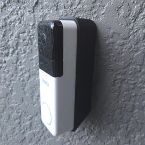 May include: A Roku smart video doorbell mounted on a textured gray wall. The doorbell has a black top section with a camera lens, a white central section with a circular button, and a black mounting bracket. The Roku logo is visible.