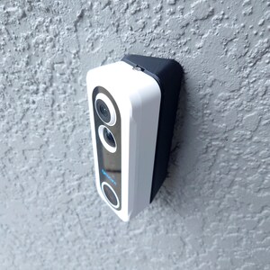 Puede incluir: Un timbre inteligente blanco y negro con cámara y logotipo azul, montado en una pared gris texturizada. El timbre tiene un diseño elegante y moderno con una parte superior redondeada y un cuerpo rectangular.