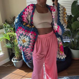 May include: A colorful, handmade crochet cardigan with a granny square design. The cardigan features a mix of pink, blue, yellow, and white squares. Paired with pink corduroy pants and a beige crop top.