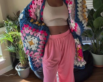 Puede incluir: Un cárdigan de crochet hecho a mano con un diseño de cuadrados de la abuela. El cárdigan presenta una mezcla de cuadrados rosas, azules, amarillos y blancos. Combinado con pantalones de pana rosas y un top corto beige.