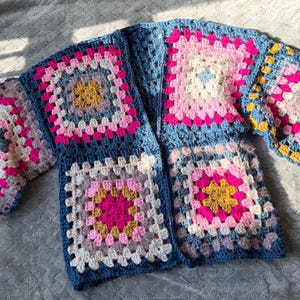 Peut inclure: Cardigan en crochet fait &agrave; la main, &agrave; motif de carr&eacute;s granny. Le cardigan pr&eacute;sente des carr&eacute;s dans les tons de bleu, rose, blanc et jaune. Les manches sont courtes et le v&ecirc;tement est pos&eacute; &agrave; plat sur une surface grise.
