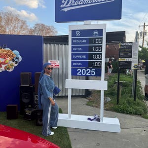 Könnte beinhalten: Ein weißes Schild mit dem Text "Dreamrulle" in blauen Buchstaben. Das Schild zeigt eine Preisliste für Benzin mit den Preisen 1,00 für Normalbenzin, Superbenzin, Super Plus und Diesel. Das Schild zeigt auch das Jahr 2025 an. Eine Person in einem blauen Jeans-Outfit steht vor dem Schild.