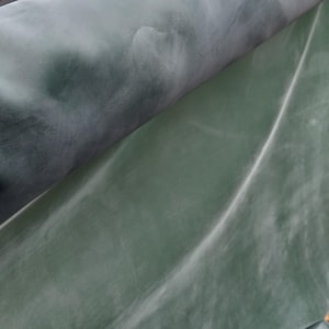 May include: A roll of smooth, flexible material featuring a gradient of dark charcoal grey transitioning into a muted sage green. The surface displays a subtle, cloudy, or marbled texture with soft, diffused light reflections across the curved form.