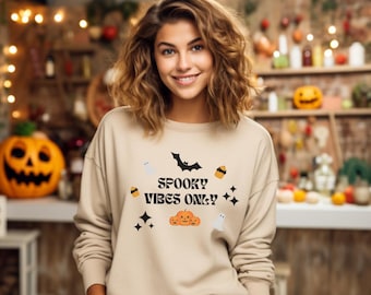 May include: Beige sweatshirt with the text "Spooky Vibes Only" and Halloween-themed graphics, including a bat, pumpkins, cupcakes, ghosts, and stars. The sweatshirt is a neutral color and suitable for the fall season.