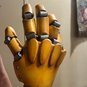 Op de afbeelding: Een gele, gelede handprop met metallic grijze gewrichten en klinknagels. De vingers en de handpalm zijn effen geel van kleur, met een licht verweerde uitstraling. De hand is ontworpen om een mechanische of robotachtige hand na te bootsen.