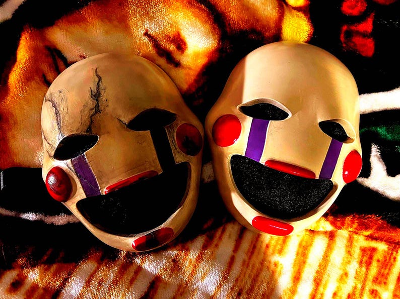 May include: Two beige-colored puppet masks with black eye sockets and mouths, red lips and cheeks, and purple accents. One mask has a cracked appearance. The masks are set against a patterned background.