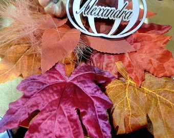 May include: A wooden pumpkin place card with the name "Alexandra" written on it. The pumpkin is surrounded by fall leaves in shades of red, orange, and yellow.